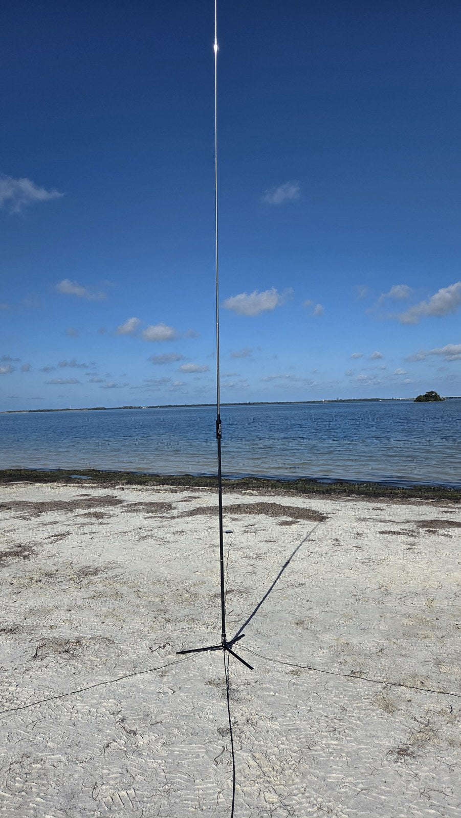 Radioddity HF-010 vertical antenna on a tripod base in the sand at Sunshine Skyway Fishing Pier State Park, with Tampa Bay and a distant island under a blue, partly cloudy sky behind it.