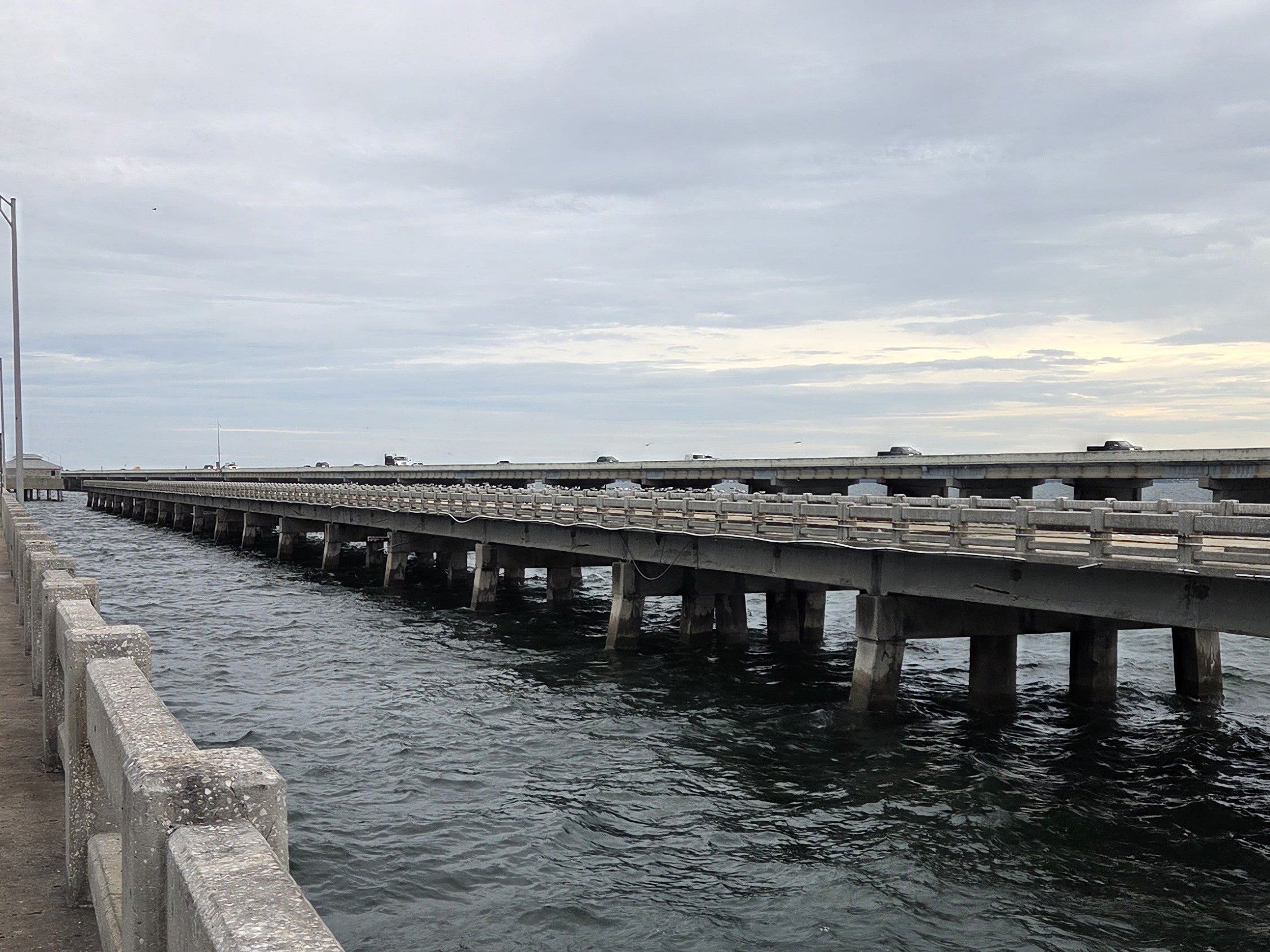 Concrete pier railing in the lower left and a long elevated roadway on concrete piers running toward the horizon over dark choppy Tampa Bay water, under a thick overcast sky with a pale yellow strip of light at the horizon.