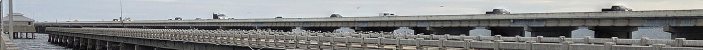 Concrete pier railing in the lower left and a long elevated roadway on concrete piers running toward the horizon over dark choppy Tampa Bay water, under a thick overcast sky with a pale yellow strip of light at the horizon.