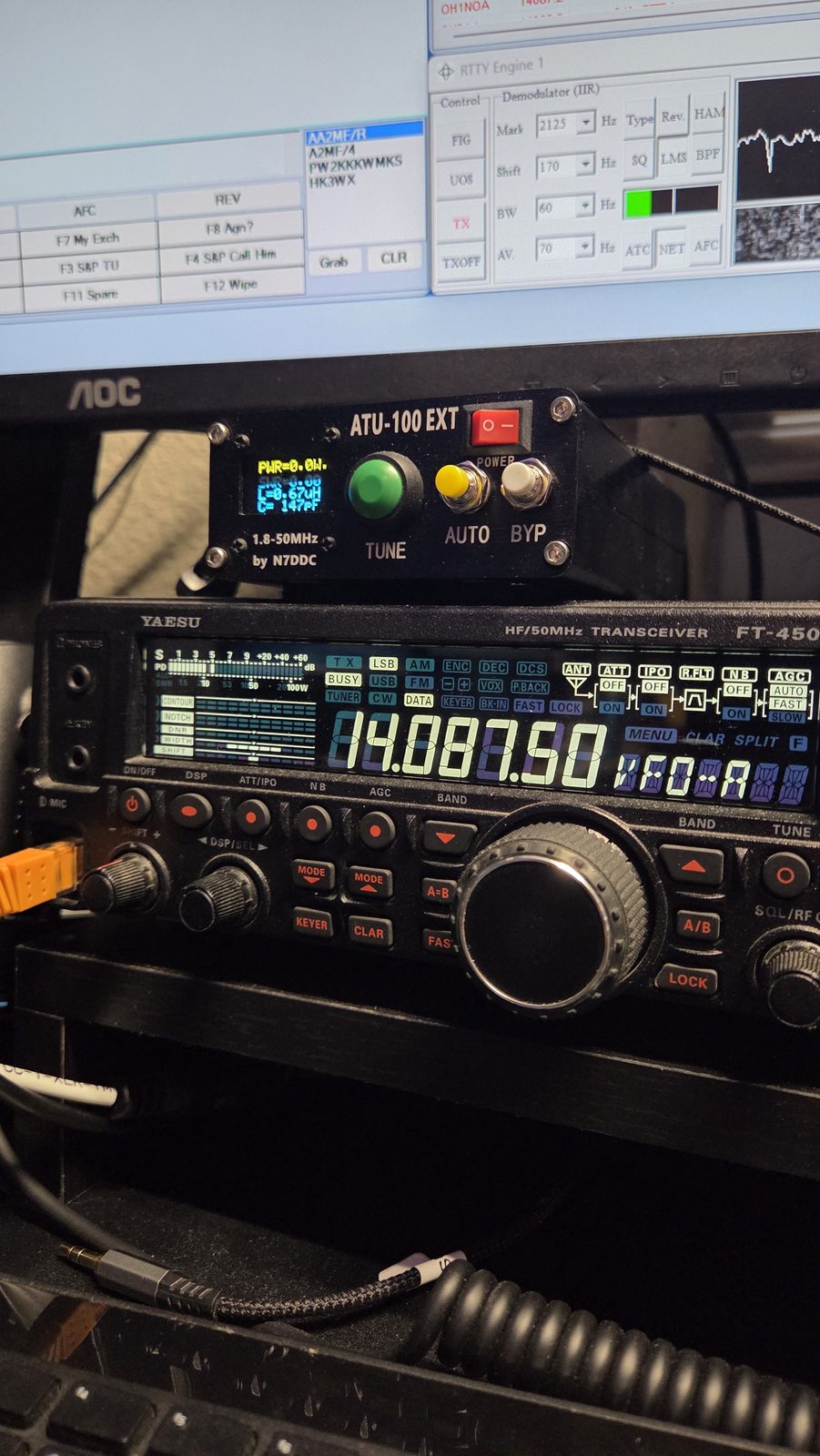 Yaesu FT-450 transceiver tuned to 14.087.50 MHz with an N7DDC ATU-100 EXT antenna tuner stacked above it, N1MM Logger+ and MMTTY visible on the monitor behind.