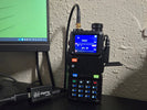 A Baofeng UV-5RM handheld standing upright on a black desk against a stucco wall, antenna cable looped overhead, display showing W4GGJ on the top line and SPA 2M below it, with a Digirig Mobile interface and cabling on the desk to the left.